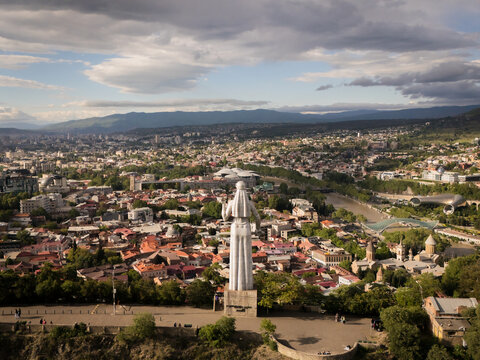 Aerial view of the Kartlis Deda statue standing tall over Tbilisi's terracotta rooftops, with distant mountains meeting the horizon, Tbilisi, Tbilisi, Georgia.