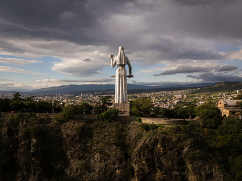 Aerial view of the majestic Kartlis Deda statue, silhouetted against the cityscape under a dramatic sky, a blend of ancient monument and modern urban sprawl, Tbilisi, Tbilisi, Georgia.