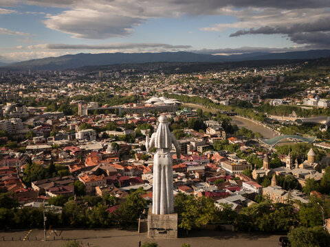 Aerial view of the Kartlis Deda statue overlooking a sprawling cityscape with terracotta roofs and distant mountains under a cloudy sky, Tbilisi, Tbilisi, Georgia.