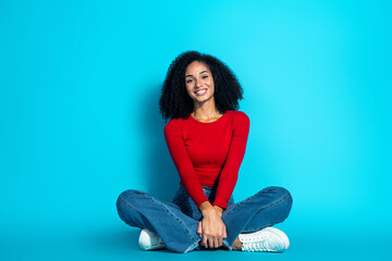 Young woman sitting with crossed legs in casual red top and jeans, smiling on a blue studio background