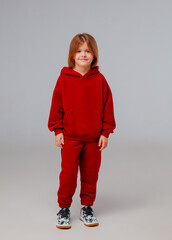 A child dressed in a red hoodie and matching sweatpants stands with a cheerful expression. The background is neutral, focusing attention on the outfit and joyful demeanor of the child.