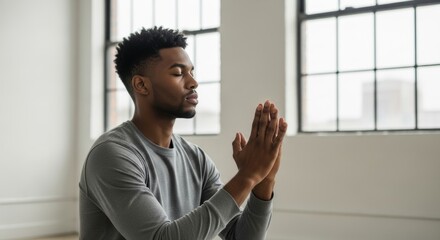 Peaceful African American man practicing meditation with hands in prayer position near large windows. Mindfulness and wellness concept in minimalist urban loft setting.