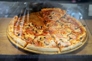 Pizza with mushrooms and ham. Freshly baked pizza with mushrooms and ham on wooden tray in window of pizzeria, warm lighting. Concept of fast food, takeaway option, popular fast meal