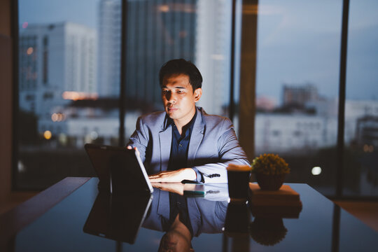 Asian businessman using tablet and working late at night in office with city lights in background, focused on work with digital device, modern workspace and technology concept