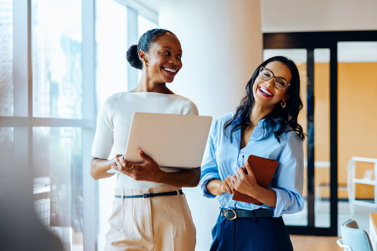 Two businesswomen smiling with a laptop and notebook in an office setting