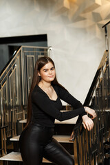 Girl in leather pants on the stairs indoors, she leans on the railing of the stairs.