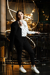 A girl at a metal railing with a large chandelier hanging behind it. She is wearing black trousers and a white blouse with a jacket.