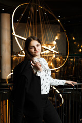 A girl at a metal railing with a large chandelier hanging behind it. She is wearing black trousers and a white blouse with a jacket.
