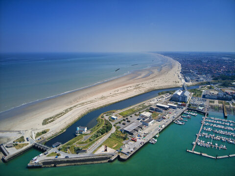 Aerial view of the shimmering turquoise sea meets the sandy beach, juxtaposed with the industrial port and cityscape, Dunkirk, Nord, France.