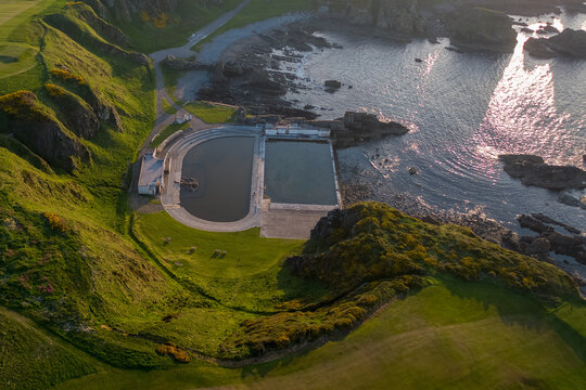 Aerial view of a concrete pool nestled between verdant cliffs and rocky shores, reflecting the sky's light, a stark contrast to the wild, rugged coastline, Macduff, Aberdeenshire, United Kingdom.