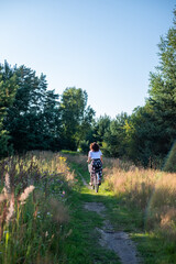 Fototapeta premium Woman riding bicycle along a scenic path through tall grass and wildflowers, surrounded by lush greenery and trees, enjoying a peaceful outdoor experience in nature