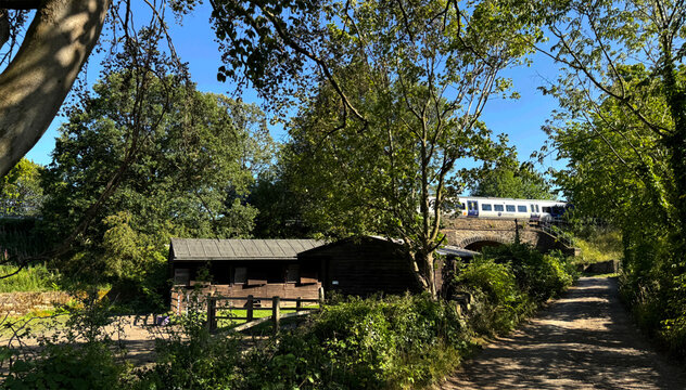 "Rustic wooden stables nestle beside a winding dirt path, framed by lush greenery. A passing train on the nearby Victorian stone bridge adds motion to the quiet charm of Norwood Green, Halifax, UK"