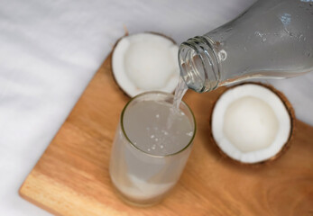 Pouring Coconut water in a glass with straw on white wooden table over white background. Fresh detox coconut juice. Selective focus.