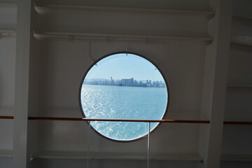 Scenic view through round porthole on cruise ship, looking out at sea and distant coastal city on sunny day