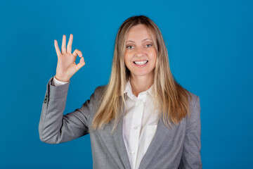 Fototapeta premium Beautiful blonde european caucasian smiling woman in business suit showing OK sign against blue background