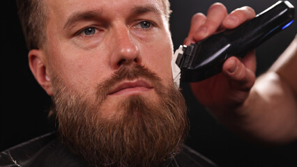 Young Man with Beard at Barber Shop. Barber is Preparing his Haircut. Dark Atmosphere, Black Background. Concept of Barber and a Client.
