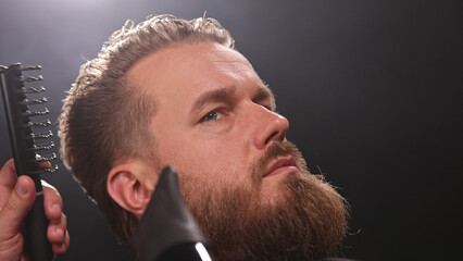 Young Man with Beard at Barber Shop. Barber is Preparing his Haircut. Dark Atmosphere, Black Background. Concept of Barber and a Client.