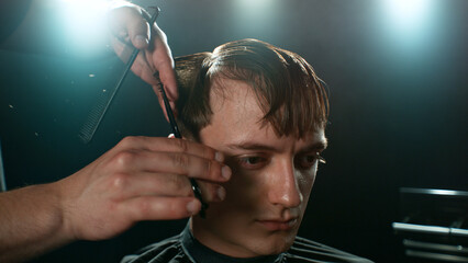 Young Man with Beard at Barber Shop. Barber is Preparing his Haircut. Dark Atmosphere, Black Background. Concept of Barber and a Client.