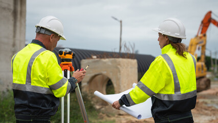 The survey team is using a theodolite and road construction plans. Civil engineers are measuring with surveying tools during the construction