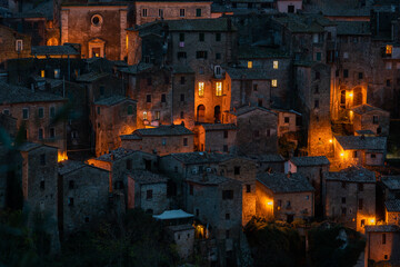 View of warm light spills from windows of ancient stone buildings, a clustered village clinging to the hillside in the twilight, Sorano, Tuscany, Italy.