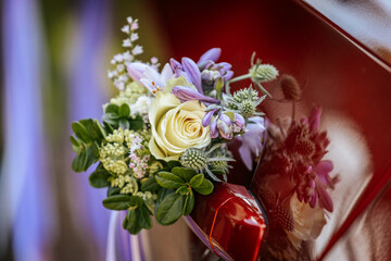 Close-up of a red wedding car door adorned with white and purple floral bouquets tied with long satin ribbons, reflecting a festive outdoor atmosphere.