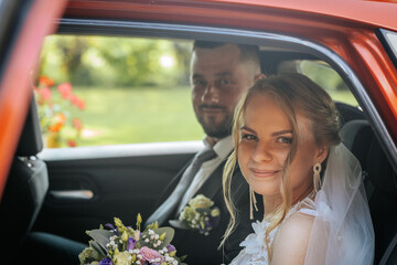 Bride and groom seated in the back of a decorated wedding car, smiling and holding a colorful bouquet. Natural light and greenery in the background.
