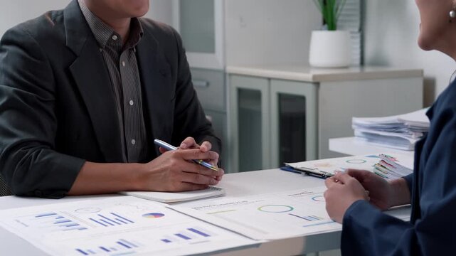 Professional business partners reviewing financial charts and graphs at office desk, shaking hands confidently after successfully reaching mutually beneficial agreement