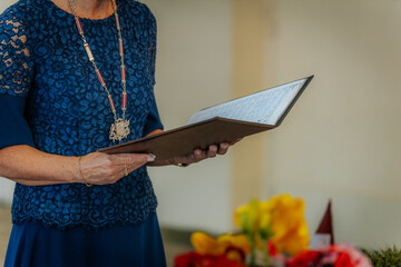 A Latvian wedding officiant in a blue lace dress holds an open marriage register, wearing a...