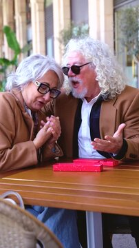 Vertical shot of senior woman receiving a red wrapped gift from her kind husband at an outdoor cafe table, both wearing matching camel coats, celebrating pure true love, surprise and enduring romance