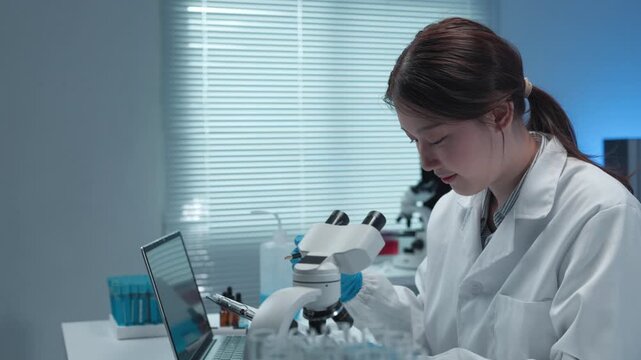 Female scientist in white lab coat analyzing medical data on laptop while working with microscope and laboratory glassware in modern sterile research environment - Powered by Adobe