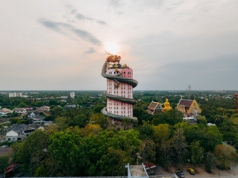 Aerial view of the striking pink temple tower embraced by a winding green dragon, sunlight glinting off its golden head, Khlong Mai, Nakhon Pathom, Thailand.