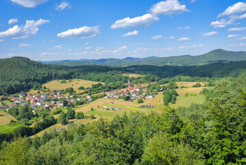 High-angle panoramic view of a small village surrounded by dense forests and rolling hills of the Pfälzerwald.