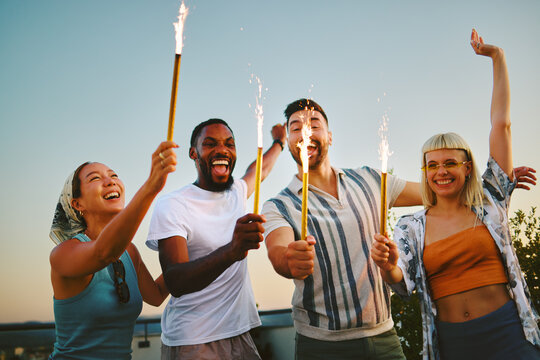 Group of cheerful multi-ethnic friends holding burning sparklers celebrating on a rooftop at sunset