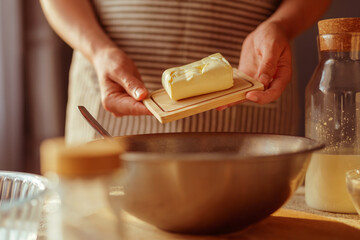 A person's hands in an apron hold a block of butter on a small wooden board, positioned above a metal mixing bowl with other baking ingredients blurred in the foreground and background.