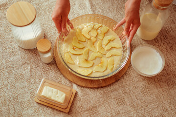 A person's hands are holding a glass pie dish filled with batter and arranged apple slices, surrounded by other baking ingredients like milk, dry goods, and butter on a patterned tablecloth.