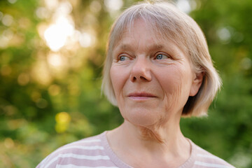 A candid outdoor portrait captures a serene middle-aged woman with short, light hair and blue eyes, looking thoughtfully to the side against a blurred, sunlit green background.