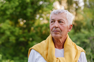 A thoughtful, outdoor portrait of a senior man with short grey hair and a yellow sweater draped over his shoulders, looking to the side against a blurred green background.