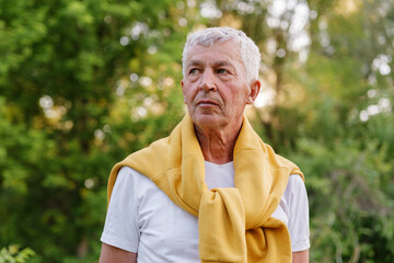 A candid outdoor portrait shows a thoughtful senior man with short grey hair and a yellow sweater draped around his neck, looking away with a pensive expression against a blurred green background.