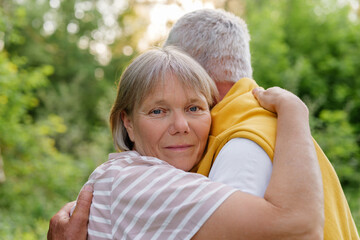 A warm outdoor portrait of a senior couple embracing, with the woman looking directly at the camera while being hugged by the man, against a backdrop of blurred green foliage and sunlight.