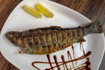 Whole grilled river trout on white plate on wooden table