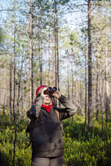 A middle-aged girl in a red hat and a green jacket stands in the forest on a sunny day and looks through binoculars into the distance.
