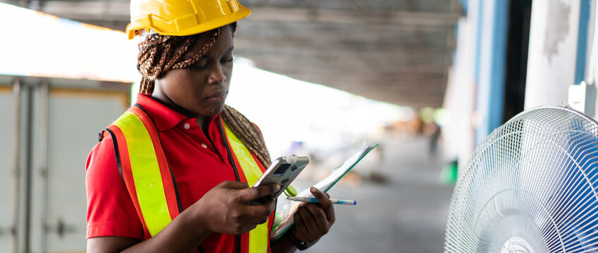 Black female industrial worker using smartphone and clipboard in a warehouse loading area. Represents mobile technology in logistics and task management