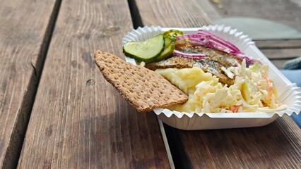 Traditional fried herring with mashed potatoes, pickled cucumbers, red onion and crispbread in a paper tray. Scandinavian street food concept with classic flavors and rustic outdoor serving