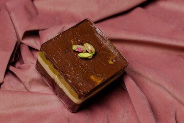 Delicious layered dessert with chocolate topping and pistachio garnish against a soft, textured pink background.