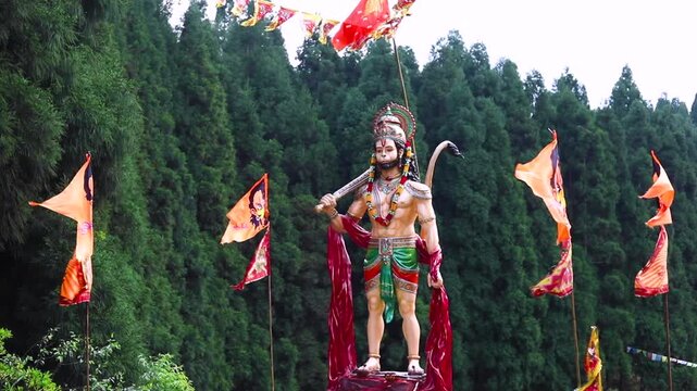 Witness the grandeur of a vibrant Hanuman statue, adorned with colorful flags, set against a lush green forest backdrop. This 10-second HD video captures the spiritual essence and beauty of the sacred