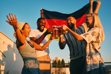 Cheerful multi-ethnic friends toasting with beer while celebrating german national holiday on a rooftop at sunset