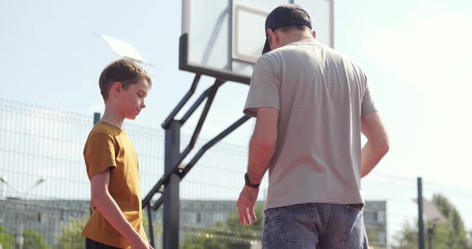 Father teaching son to throw ball into basketball hoop. Son watching father throwing ball into hoop. Family sports moment: father and son playing basketball. Family sports and fatherhood.