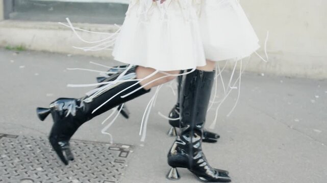 Model walking on sidewalk wearing white dress and black boots with ribbons