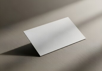 A blank, white business card rests on a textured surface, casting a soft shadow with light filtering from above.