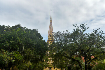 Golden Buddhist temple of Wat Chalong with intricate carvings, pointed spire, and surrounding greenery under a cloudy sky.